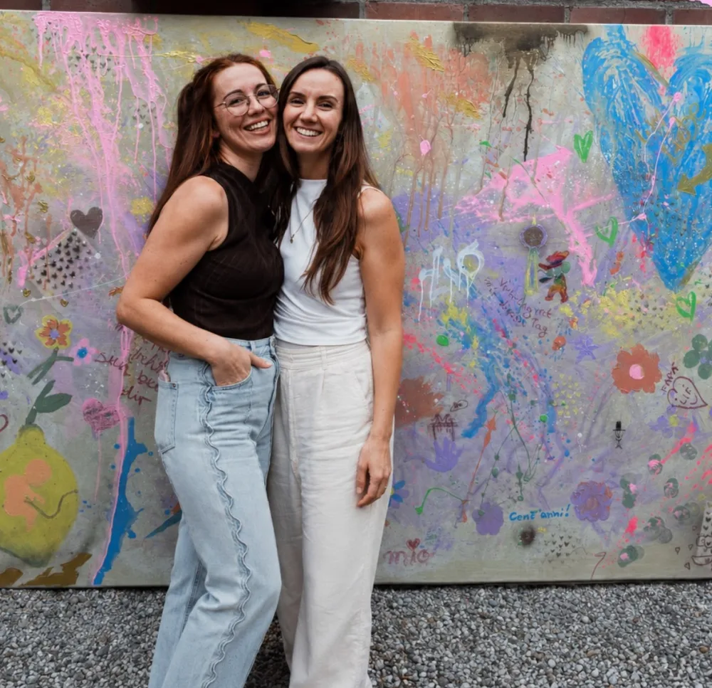 Anna-Lena und Chrissi vor einem großen Gemeinschaftsbild einer Hochzeit, auf dem sich alle Gäste mit Herzen, Blumen und Glückwünschen verewigt haben